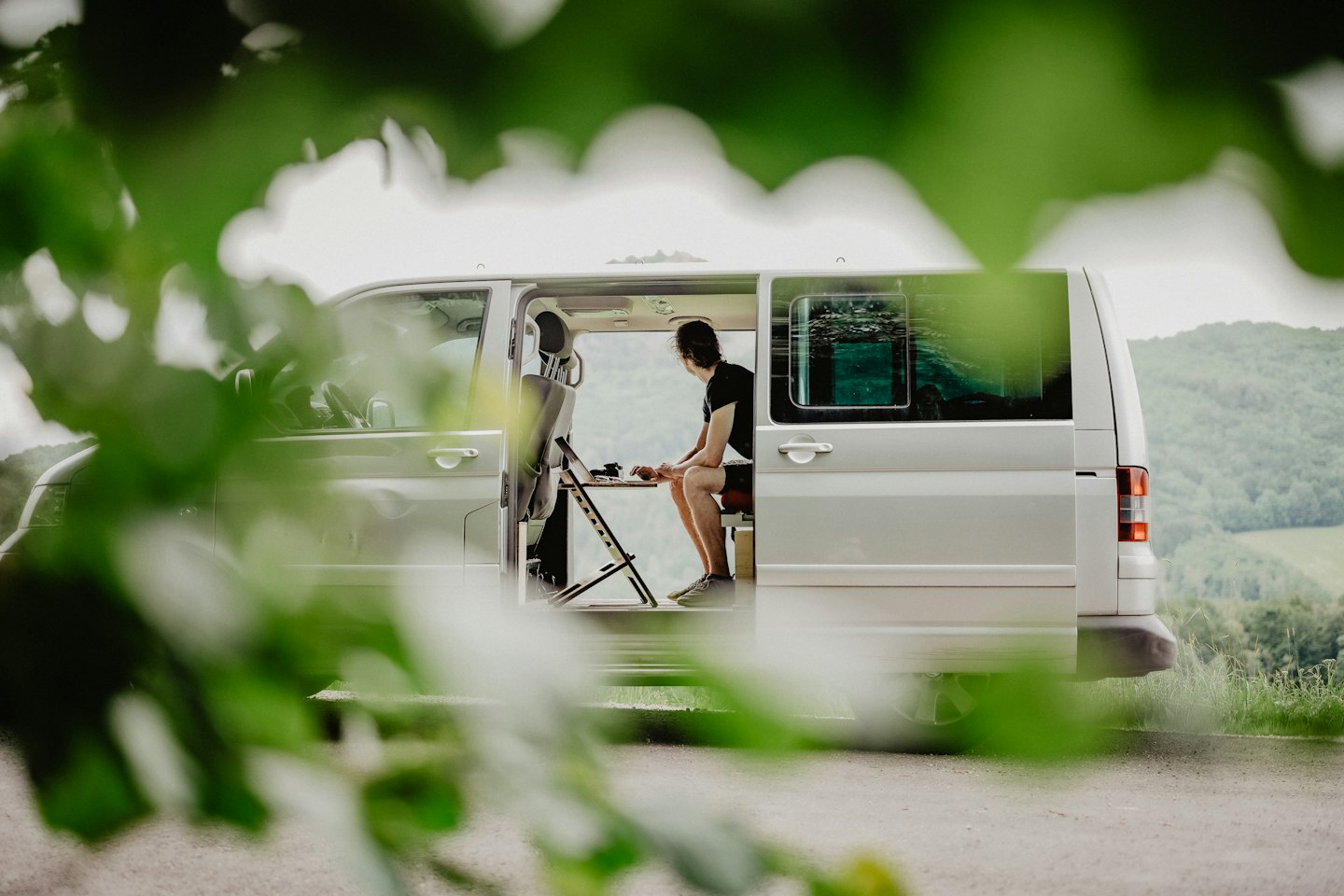 Uomo che lavora al computer portatile in un camper con colline verdi all'esterno.